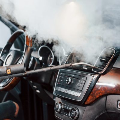 Modern black automobile get cleaned inside by steam equipment by worker inside of car wash station.